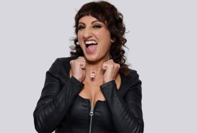 Steph Tolev joyfully in a black zip-up outfit laughs brightly, holding a small pendant before a plain gray backdrop.