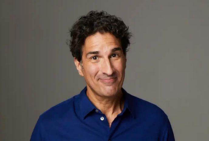 Gary Gulman smiles with a playful expression, wearing a blue button-up shirt, in gray studio background.