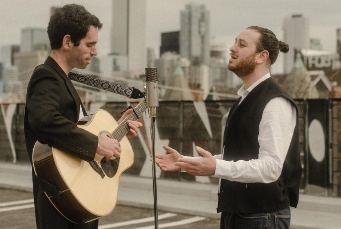 Two musicians perform on a rooftop, one playing acoustic guitar while the other sings passionately into a microphone.