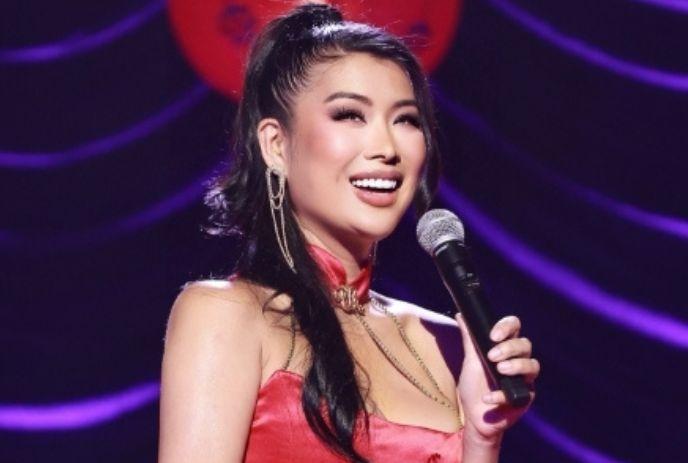 Smiling performer in a red satin dress holding a microphone on stage, with elegant earrings and purple lighting
