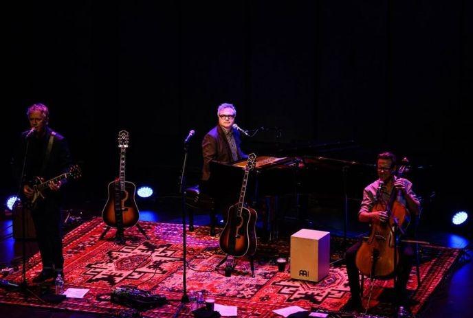 Musicians perform on a dimly lit stage with guitars, piano, and cello, creating an intimate live concert scene.