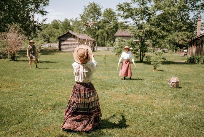 Three people in vintage clothes play with hoop on grass near rustic wooden houses and trees under soft daylight.