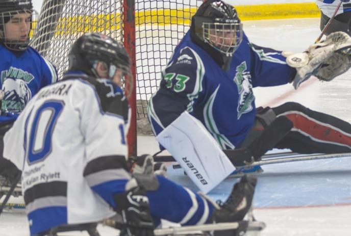 Sled hockey goalie in blue jersey defends net as white jersey player approaches, puck and rink lines visible.