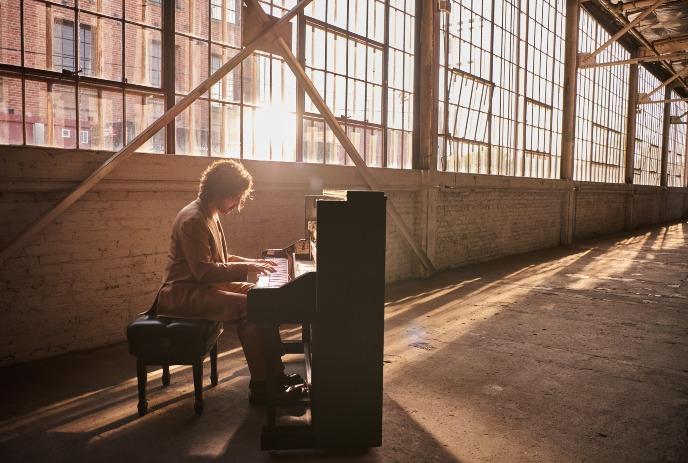 Person in brown suit playing upright piano in sunlit industrial room with large windows, brick walls, and warm ambiance.