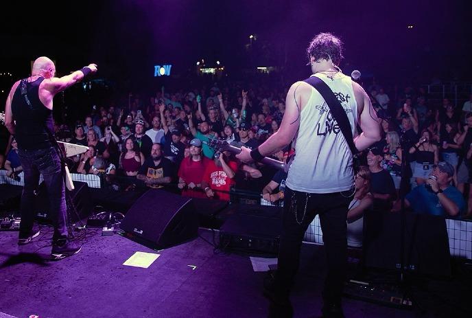 Two guitarists perform onstage as a cheering crowd raises hands under bright purple lights.