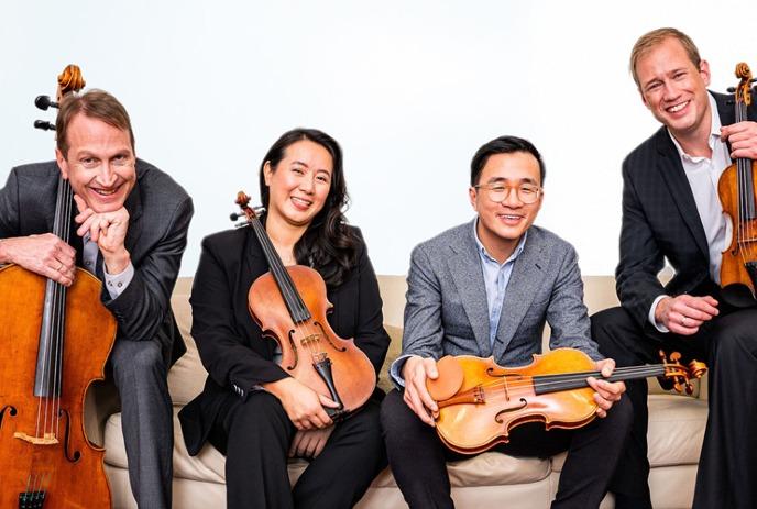 Four musicians seated on a couch holding string instruments—two cellos, a viola, and a violin—in formal attire.