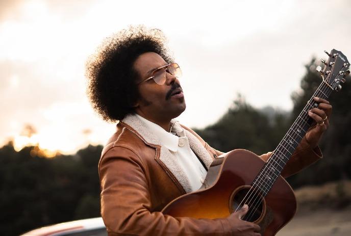 Alex Cuba playing an acoustic guitar with a sunset in the background, wearing sunglasses and a jacket.