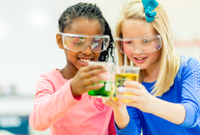 Two girls with safety glasses on looking at measuring cups with green and yellow water.