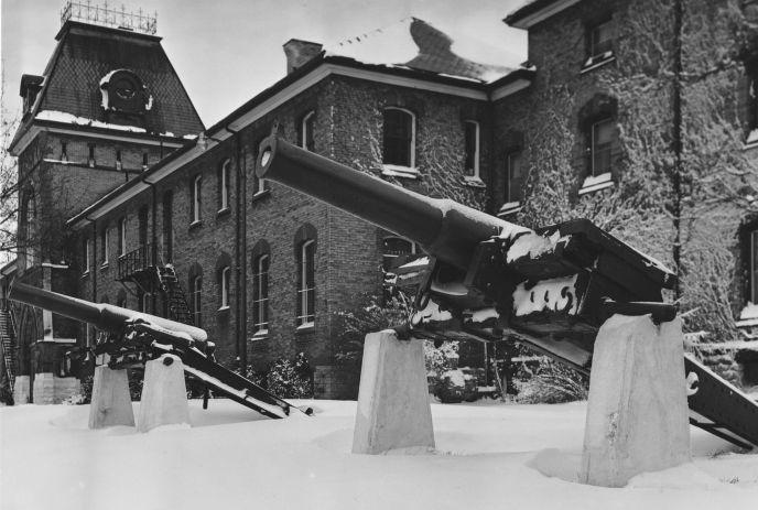 Two large cannons sit on snowy concrete before tall brick building with arched windows and clock tower.
