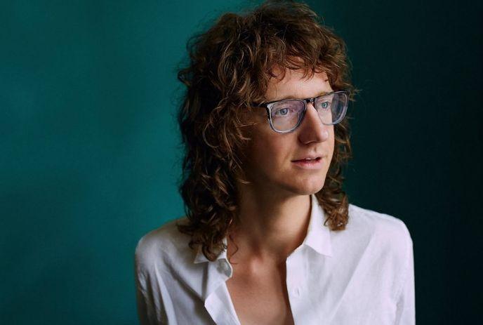Chris Fleming, with curly dark hair and glasses, wears a white shirt, gazing thoughtfully off-camera against teal background.
