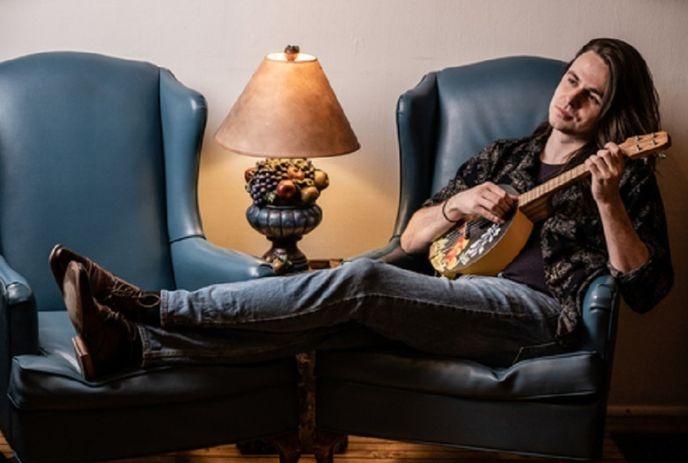 Stephen Ingram lounging across two blue chairs, playing a banjo ukulele, beside a warm lamp, looking relaxed.
