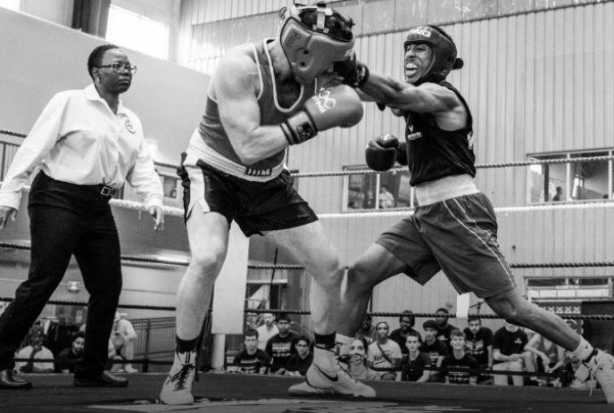 Two boxers in headgear spar in a ring while a referee and spectators watch in a black-and-white scene.