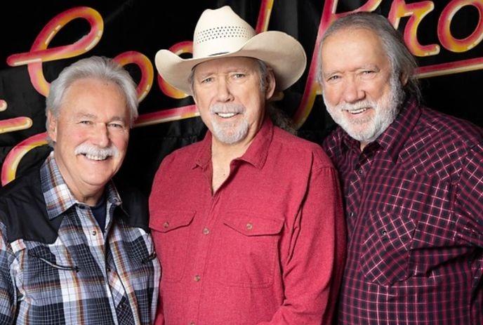 Three men together in a line, smiling, one in a cowboy hat, wearing casual shirts in front of a dark backdrop.