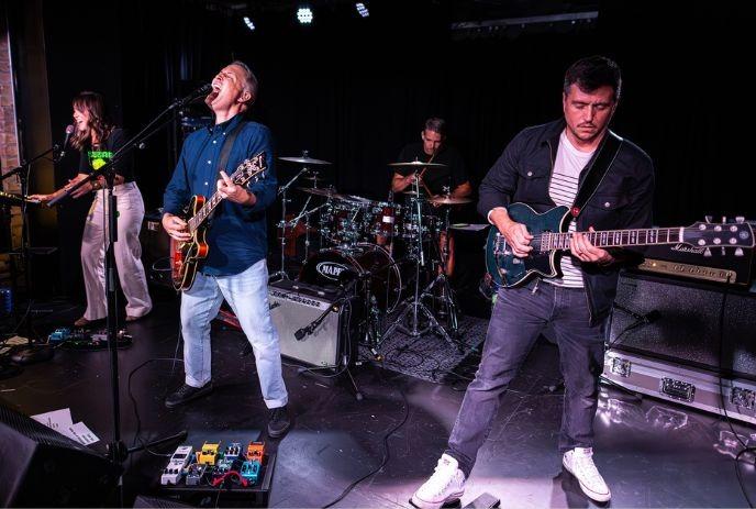 Four band members performing on the stage as singer, guitarist, drummer and keyboardist play under spotlight.