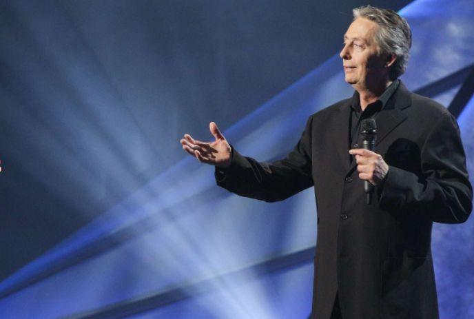 A speaker in a black suit holds a microphone and gestures on a blue-lit stage with dramatic light beams in the background.