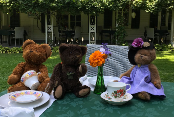 Three teddy bears sit on a lawn with plates and tea cups and an orange flower in a vase.