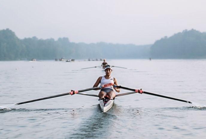 Rower glides across Fanshawe Lake, with other rowing boats, misty trees, calm water and a hazy morning sky.
