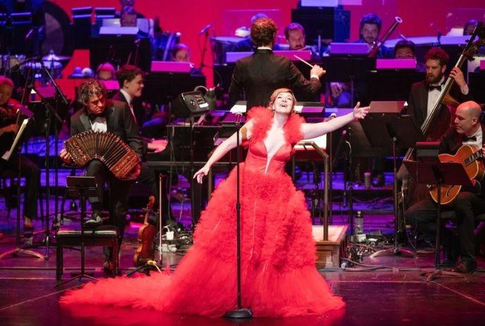 Singer in a vivid red gown performs before an orchestra as the conductor leads a vibrant concert.