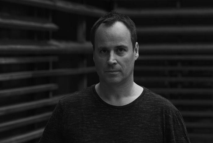 A black-and-white portrait of Christian Smith looking forward against a structured, dark slatted wall.