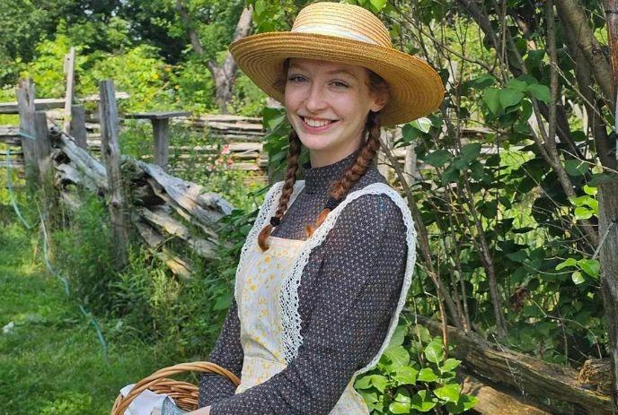 Girl dressed as Anne of Green Gables, holding a basket outdoors at Fanshawe Pioneer Village, smiling in the sunshine.