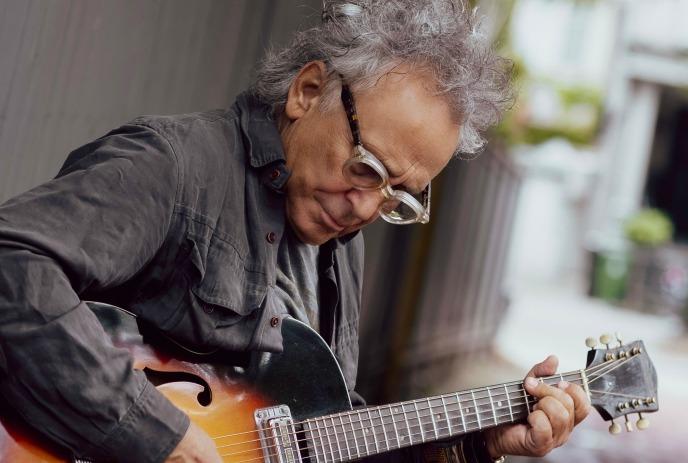 Marc Jordan with gray hair plays an acoustic guitar. His expression is pensive, evoking a soulful, creative mood.