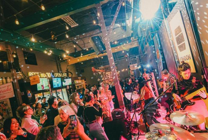 Band performing on lit indoor stage as crowd records with phones in warm venue decorated with signs and lights.