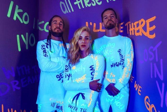 Walk off the Earth members posing in glowing white outfits with colorful text against a vibrant, graffiti-covered purple wall.