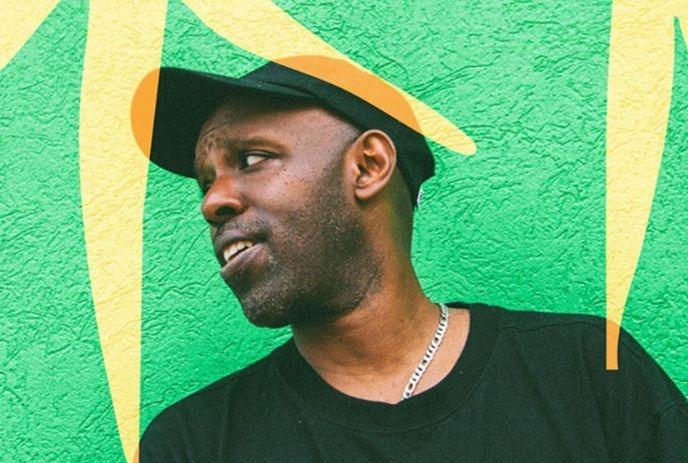 Shad wearing a black shirt and black hat, wearing a silver chain and standing against a green wall.