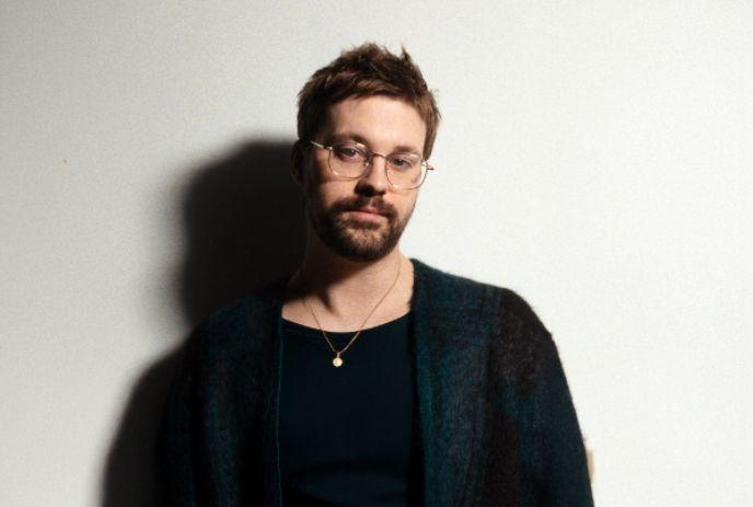 Derik John Baker wearing a sweater and glasses, standing in front of a white background.