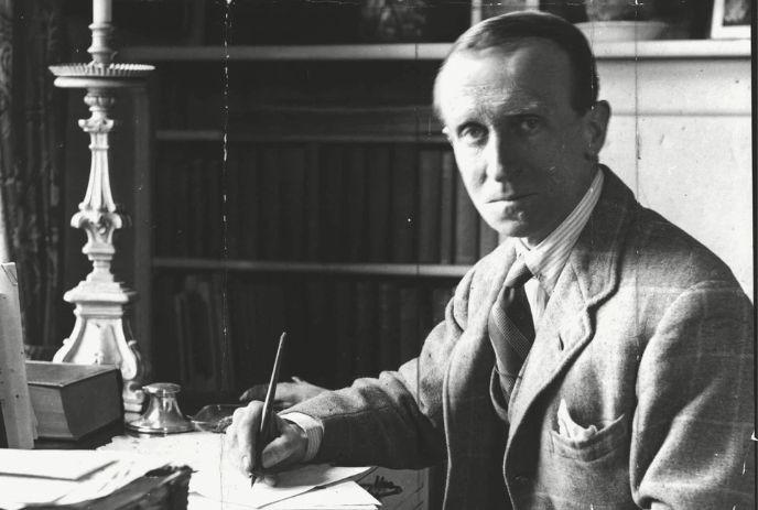 A black and white photo of John Buchan, sitting at a desk with a pen in his hand.
