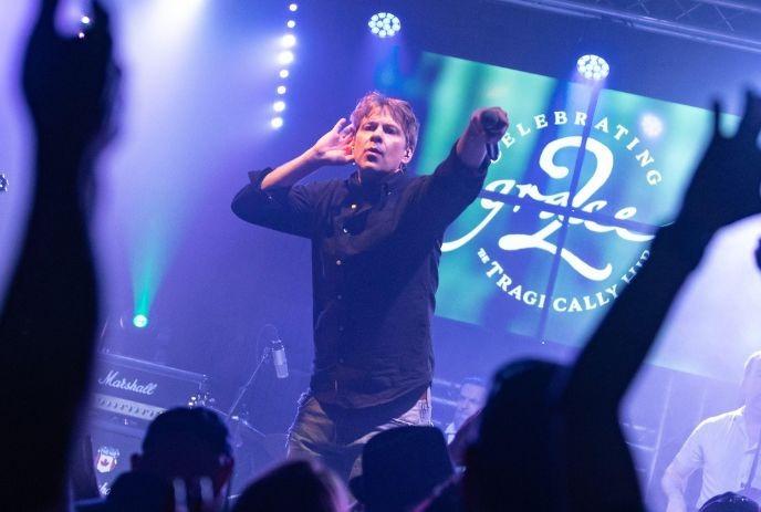 A man on stage holding a microphone out to a crowd with a hand up to his ear.