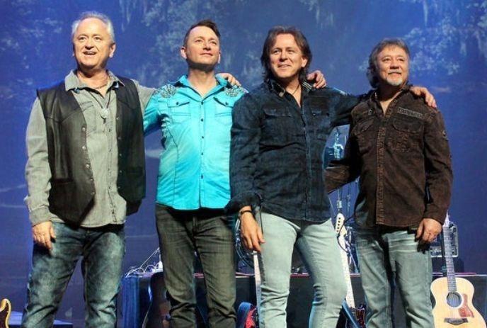 The 4 band members of Hotel California with their arms around each other, looking out at an audience while on stage.
