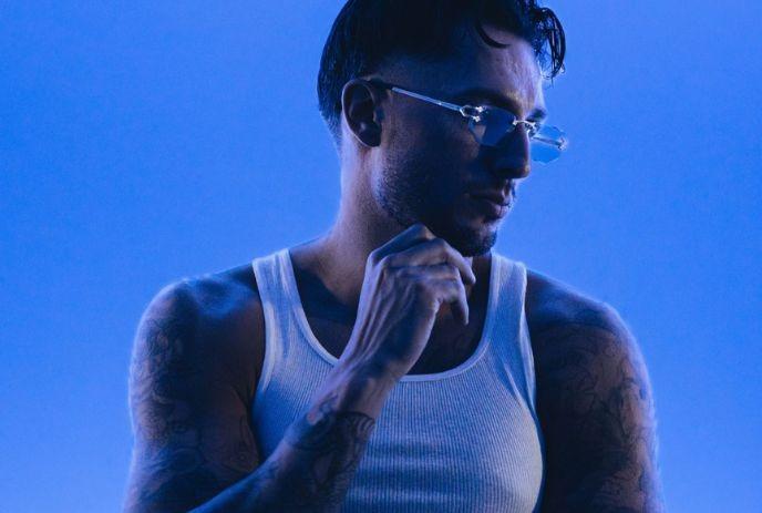 BIJOU wearing a white tank top and glasses, posing for the camera in front of a blue background.