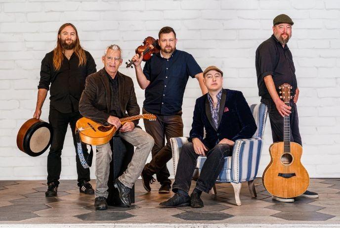 The Steel City Rovers pose against a white brick wall. They hold various instruments, conveying a relaxed, artistic vibe.