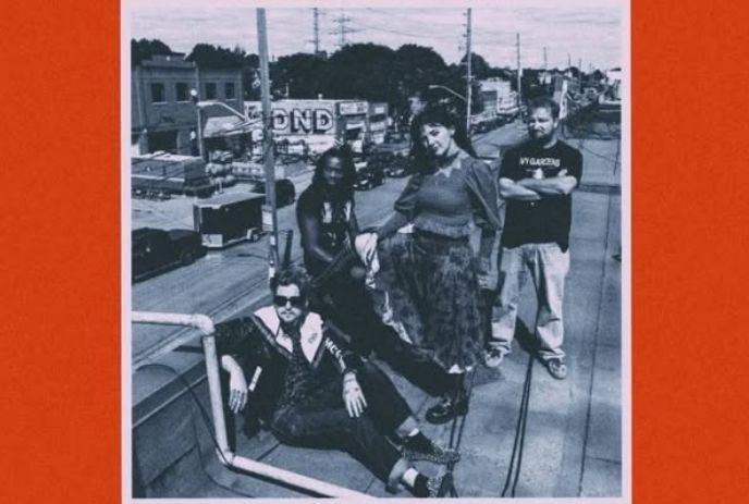 A group of four people stand and sit on a rooftop, overlooking a cityscape with street signs and buildings.