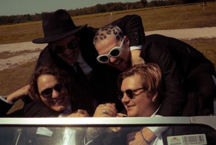 The four band members of Hartlet in suits gathered around a convertible's front seat, laughing and wearing sunglasses.