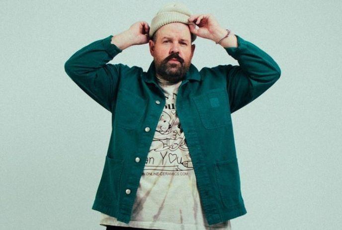 Donovan Woods in a green jacket and graphic t-shirt adjusts his beige beanie.