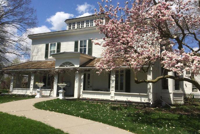 Eldon House with a pitched roof and large porch, surrounded by a lush green lawn and a blossoming cherry tree.