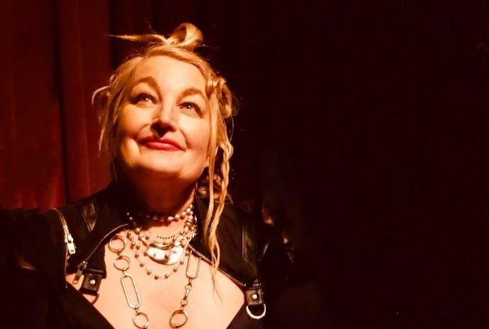 Jane Siberry with a radiant smile and blonde hair styled in an updo, wearing layered necklaces and a black outfit.