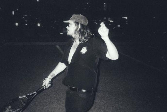 Brett Emmons with long hair and a cap swings a tennis racket energetically at night.