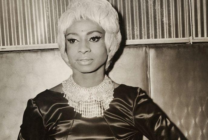 Jackie Shane with a blonde beehive hairstyle, wearing a dark satin dress and a sparkling statement necklace.