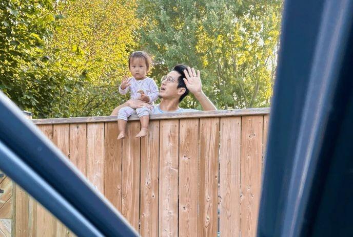 A person waves while holding a seated child on a wooden fence, with trees in the background.