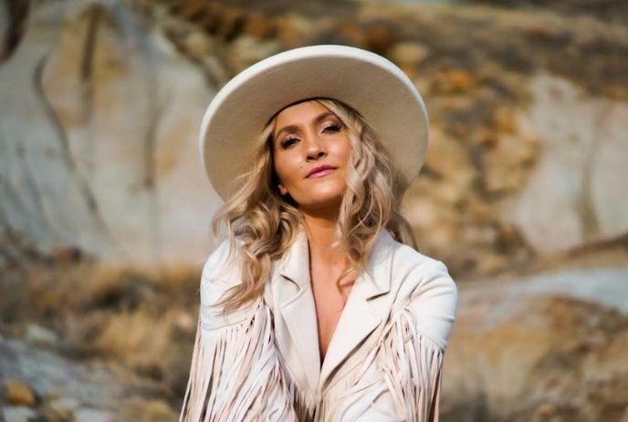 Kalsey Kulyk with long, wavy blonde hair wears a white fringed jacket and wide-brimmed hat against a rocky desert backdrop.