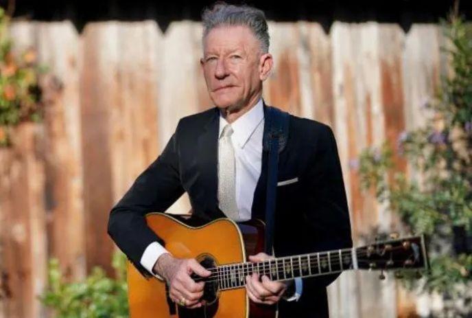 Lyle Lovett standing outside in front of a wooden fence, wearing a suit and holding a guitar.