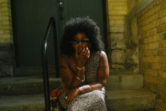 Fiker sitting on steps in front of a brick building, wearing a sleeveless dress and sunglasses.