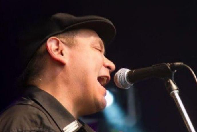 Robbie Antone wearing a black hat, singing into a microphone on stage.
