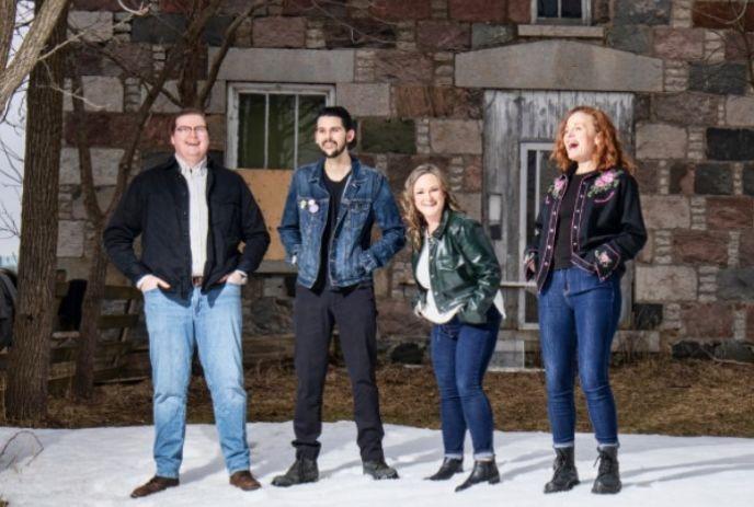 A photo of the band Maggie's Wake, featuring four members standing in front of a stone building with snow on the ground.