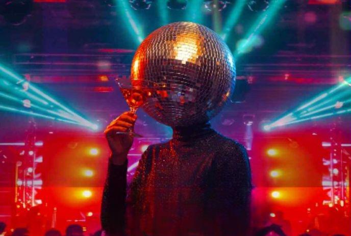 A person with a disco ball head holds a cocktail glass in a vibrant nightclub with colorful lights.