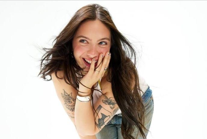 Saveria laughing with hands near her face, showing tattooed arms and casual attire.