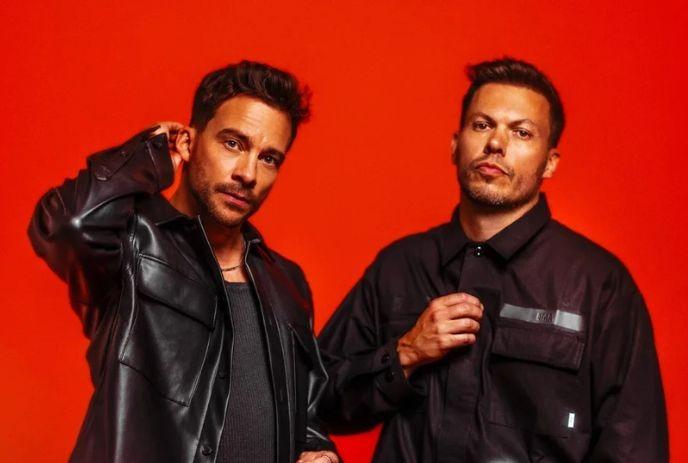 The two band members of Delta Heavy posing in front of the camera wearing black coats, in front of a red background.